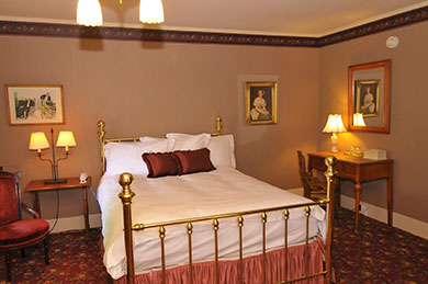 hotel room with queen bed that has brass bars surrounded by two tables lamps and victorian theme pictures on wall