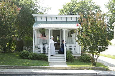 two women dressed up in victorian dresses on front porch of small white cottage surrounded by trees on a sunny day